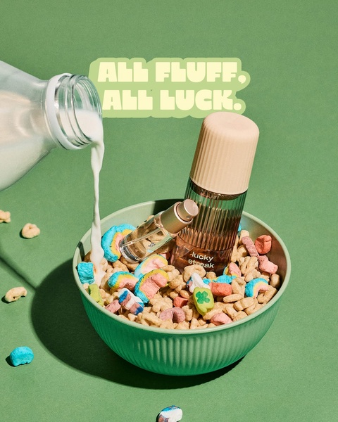 A perfume bottle is placed inside a bowl of colorful, marshmallow-filled cereal while milk is poured, creating a playful and high-contrast visual.