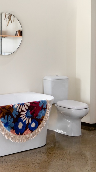 A high-contrast 9:16 shot featuring a circular, floral-patterned bath mat placed against a stark white bathroom interior to highlight design and texture.