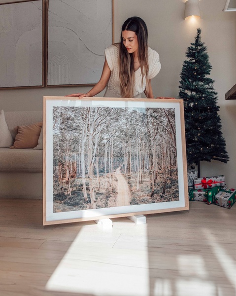 A woman stands behind a large framed landscape photo, showcasing it as a premium home decor item within a decorated, holiday-themed living room space.