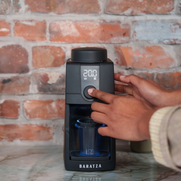 A close-up shot showing a hand interacting with the control dial of a black Baratza coffee grinder against a warm, rustic brick background.