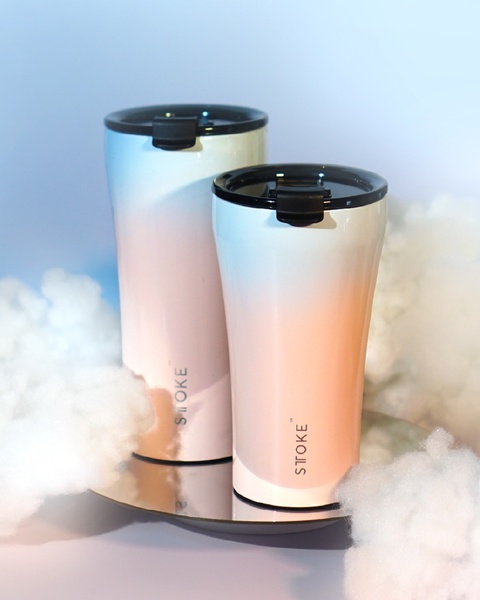 A high-end, clean product shot of two gradient-colored tumblers resting on a reflective mirror surface surrounded by soft, white clouds.