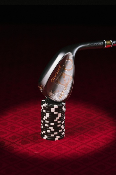 A premium golf wedge is perfectly balanced on a stack of black casino chips, set against a deep red textured background to evoke a sense of risk and precision.