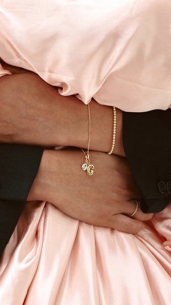 A close-up, elegant shot of a person wearing a delicate gold initial necklace and matching bracelet, framed in soft pink fabric.