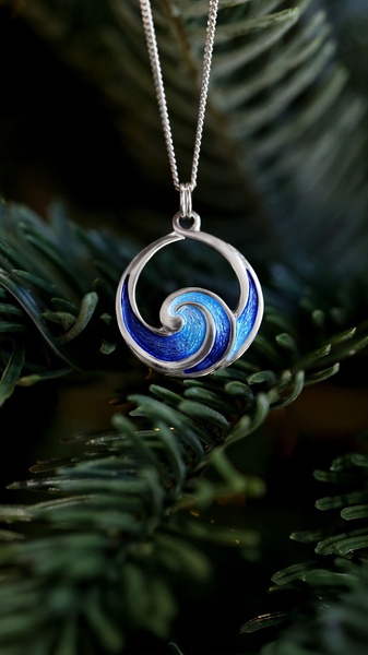 A high-quality close-up of a silver wave pendant necklace resting against deep green pine needles to evoke a seasonal, premium feel.