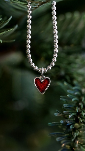 A close-up vertical shot of a silver beaded necklace with a red heart pendant hanging on a festive evergreen branch.