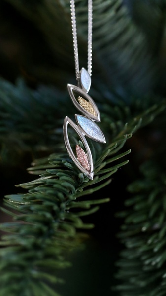 A close-up vertical hero shot of a delicate, multi-toned leaf-inspired necklace resting on pine branches.