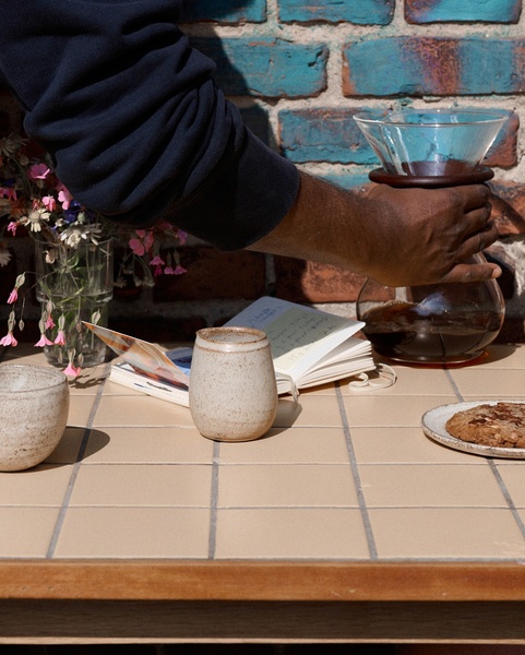 A high-quality lifestyle shot featuring a glass coffee carafe being handled on a tiled table, emphasizing a calm and ritualistic atmosphere.