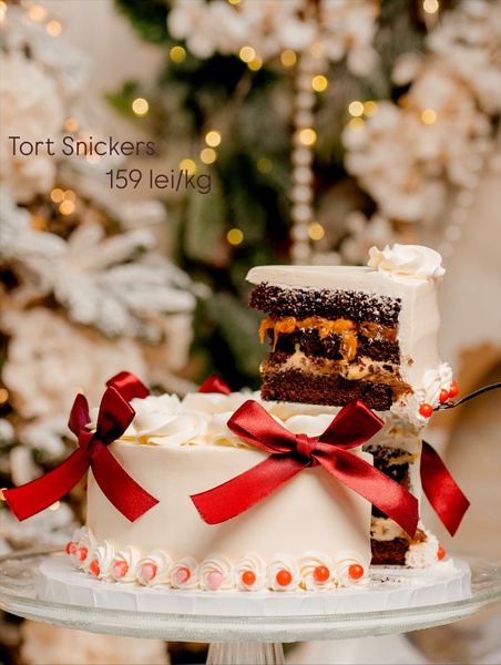A high-end, studio-lit photograph of a Snickers cake served on a glass stand with holiday-themed decorations and red ribbon accents.