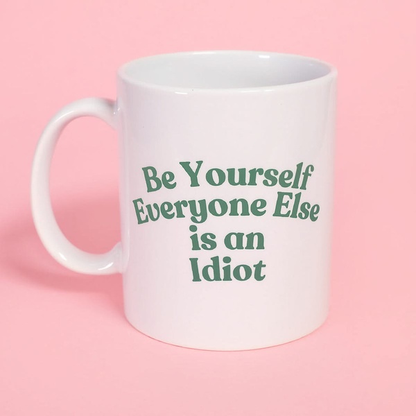 A static 1:1 image featuring a white ceramic coffee mug with the text 'Be Yourself Everyone Else is an Idiot' against a solid pastel pink background.