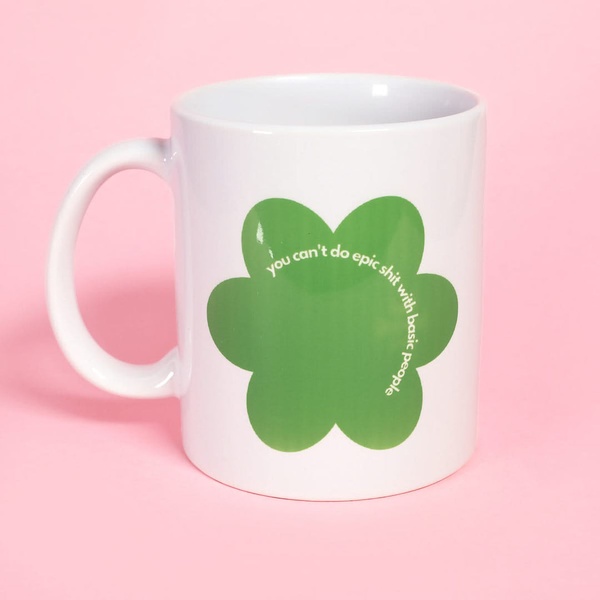 A 1:1 square static ad showcasing a white ceramic mug centered on a solid pastel pink background, featuring a green flower icon with bold white text.