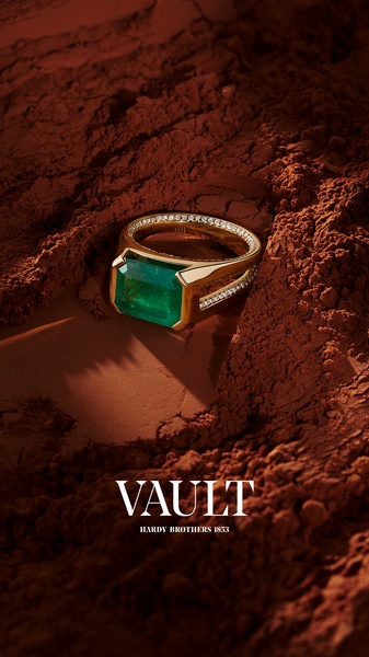 A high-end jewelry advertisement featuring a gold emerald ring set within deep, rustic earth-toned sediment to create a luxury contrast.