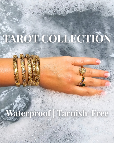 A close-up of a hand wearing gold bangles and rings submerged in bubbling water, highlighting the waterproof and tarnish-free features of the collection.