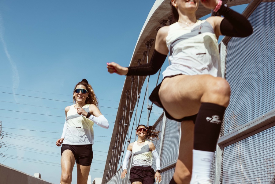 A low-angle, high-energy shot of three runners wearing matching athletic gear, specifically highlighting running arm sleeves in a bright, outdoor urban environment.