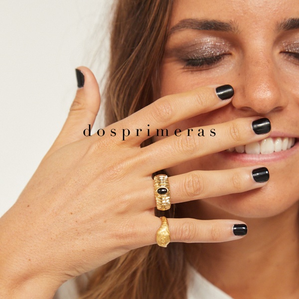 A close-up shot of a hand wearing two gold rings with a neutral white background and black nail polish aesthetic.