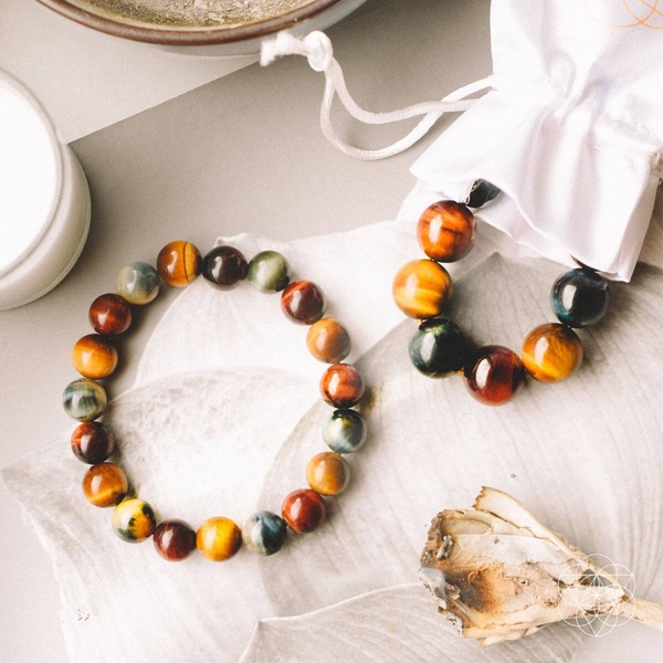 A high-quality still life image featuring multi-colored gemstone bracelets displayed next to a white pouch and dried botanical elements.