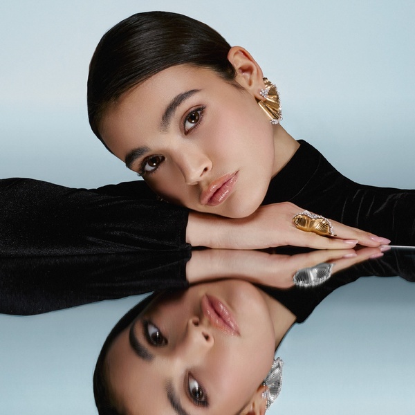 A high-fashion, minimalist close-up of a model wearing elegant gold earrings, captured with a reflective surface and cool-toned lighting.