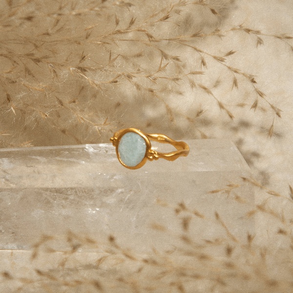 A close-up product photograph of a gold ring featuring a light blue stone, placed on a transparent crystal block against a soft textured background.