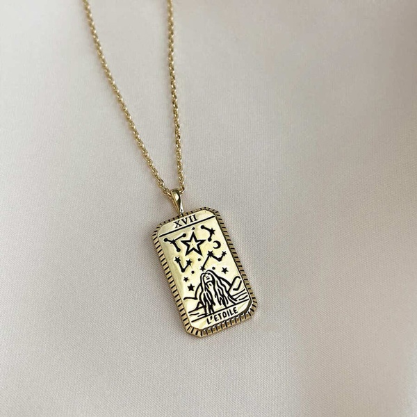 A close-up photograph of a gold-toned rectangular pendant necklace featuring a tarot card engraving, showcased against a soft, neutral fabric background.