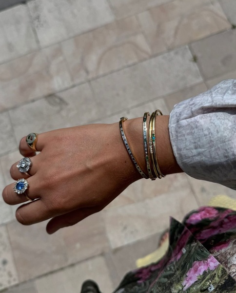 A close-up, natural-light shot showcasing a collection of three stacked gold bracelets on a wrist, paired with classic ring accents.