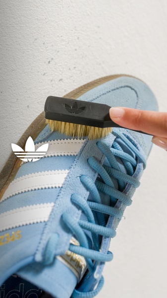 A focused, high-contrast 9:16 vertical shot showing a hand using a specialized brush to clean the texture of a light blue suede sneaker.