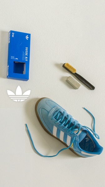 A high-contrast, overhead product layout featuring a suede sneaker, cleaning kit box, and brush set against a clean, neutral background.