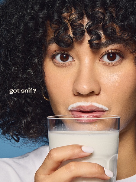 A close-up portrait of a model holding a glass of milk with a milk mustache, subverting the classic campaign imagery for a perfume brand.