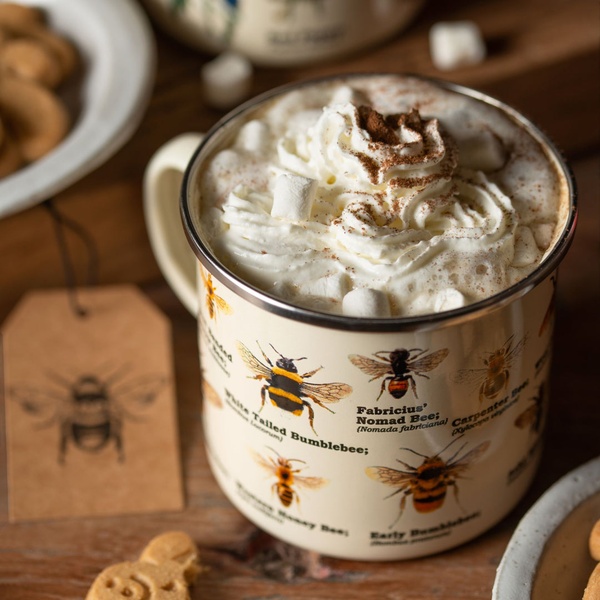 A close-up, high-quality shot of a ceramic mug filled with hot cocoa and whipped cream, featuring detailed illustrations of various bees, set in a cozy, rustic environment.