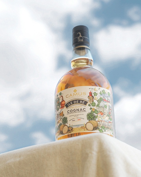A high-quality studio-style product shot of a cognac bottle placed on a simple cloth surface against a clear blue, cloudy sky backdrop.
