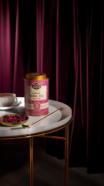 A high-end, dramatic still life photograph of a tea canister on a minimalist table, set against rich, deep purple curtain drapes to evoke a luxury experience.