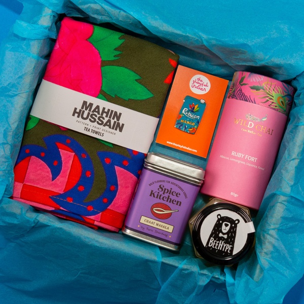 A high-angle flat-lay shot of a curated gift box featuring tea, spice mix, a kitchen towel, and a collectible pin.