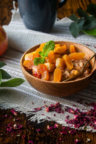 A close-up, high-quality food photography shot of an oatmeal bowl topped with dried apricots and fresh mint, set on a rustic textile background.