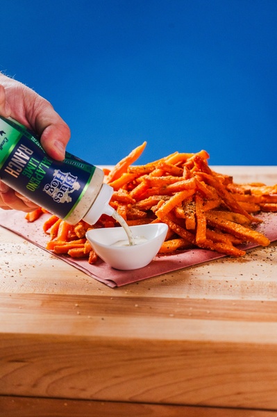 A close-up action shot showing dipping sauce being poured over a pile of seasoned fries. The vibrant blue background creates high contrast with the orange food.