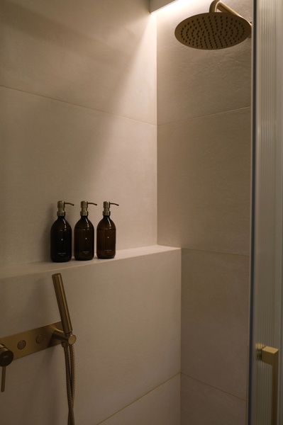 A close-up, high-end photograph of matching shower dispensers in a modern bathroom, featuring a warm, neutral color palette and brass accents.