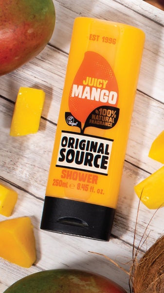 A close-up studio shot of a mango-scented shower gel bottle surrounded by fresh mango slices and coconut textures on a rustic wood surface.