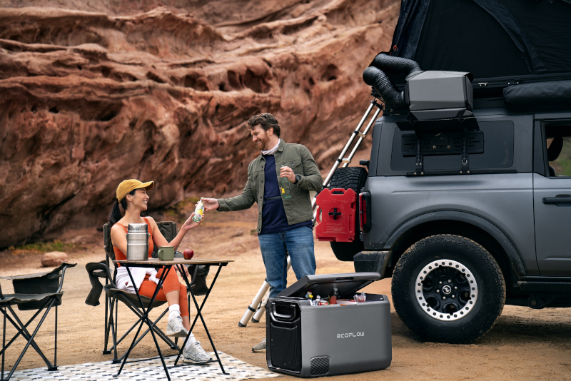 A high-quality lifestyle shot featuring a portable cooler next to a modern off-road vehicle in a rugged canyon setting.