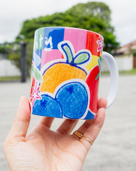 A close-up photograph of a brightly decorated ceramic mug featuring fruit illustrations, held by a hand to provide a sense of scale and real-world usage.