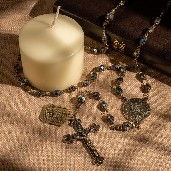 A high-quality studio shot of a metal rosary laid across a burlap surface, positioned next to a closed Holy Bible and a large pillar candle.