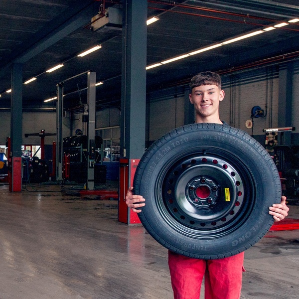 A smiling mechanic in a bright workshop setting holds a spare tire, highlighting product accessibility and professional service.