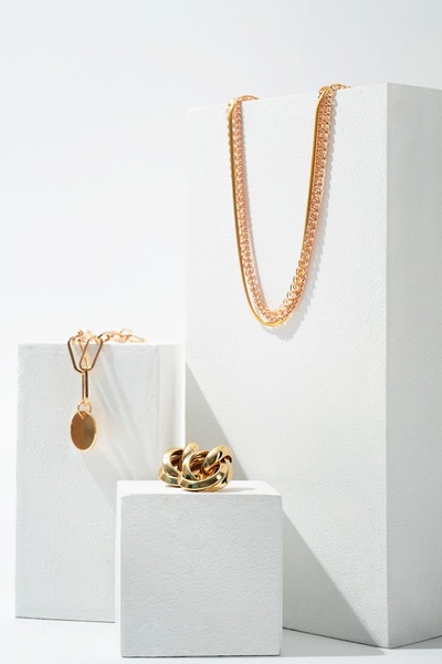 A high-end, clean studio product shot featuring gold chains and earrings displayed on white geometric blocks.