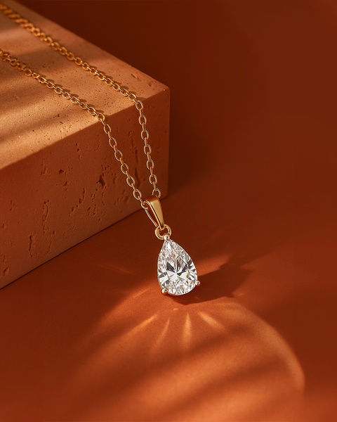 A close-up aesthetic shot of a pear-cut diamond pendant necklace resting on a terracotta block, illuminated by dramatic sunset-style lighting.