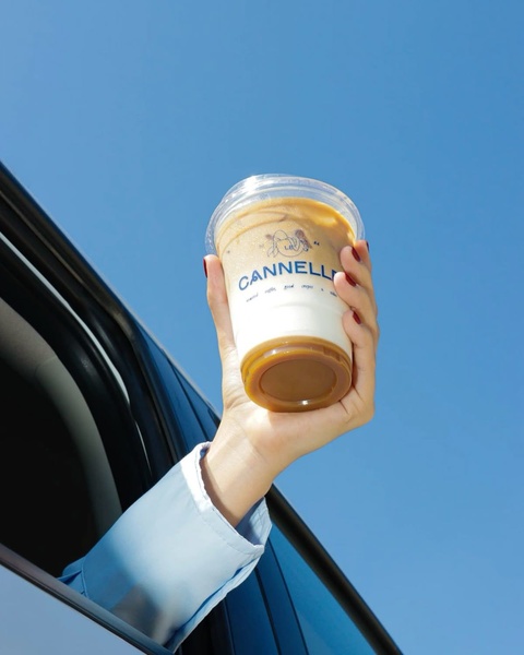 A first-person point of view shot holding a branded iced coffee cup out of a car window against a clear blue sky.