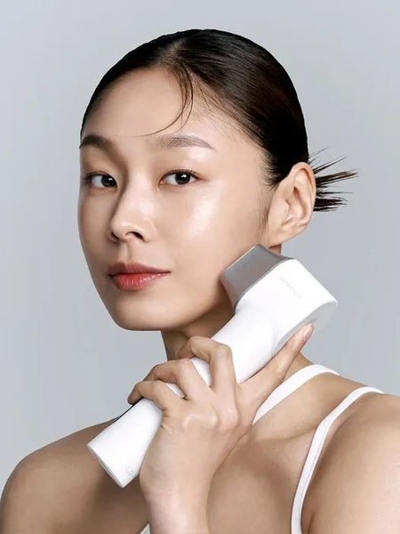 A clean, high-key studio shot of a model using a handheld facial massager device to emphasize skin texture and product application.