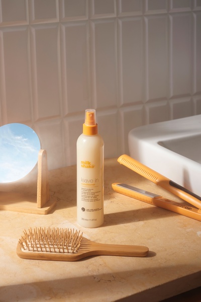 A high-quality product hero shot featuring a leave-in conditioner spray bottle centered on a bathroom vanity with hair styling tools.