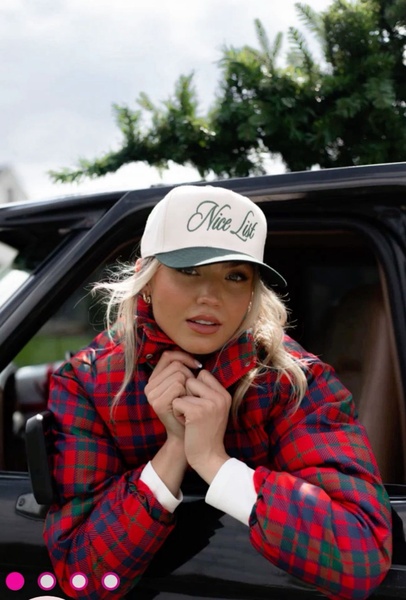 A high-quality lifestyle shot of a woman wearing a 'Nice List' trucker hat, leaning out of a vehicle in a festive, outdoor winter setting.
