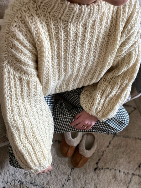 A close-up, top-down shot of a thick, off-white ribbed knitted sweater paired with casual checked pants and slip-on loungewear.