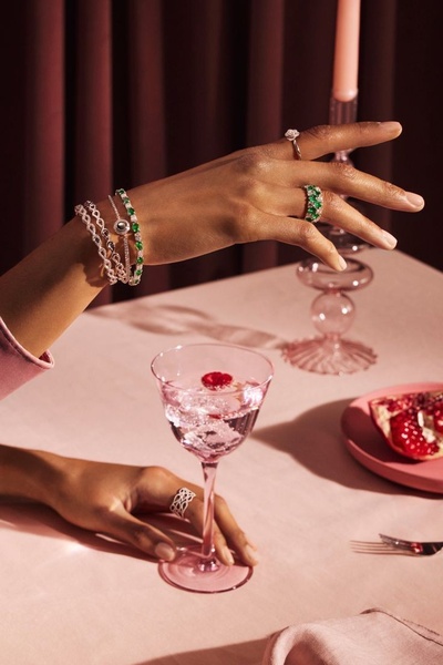 A close-up, atmospheric lifestyle shot featuring a model's hand adorned with luxury rings and bracelets, posed gracefully over a table with a cocktail and fruit.