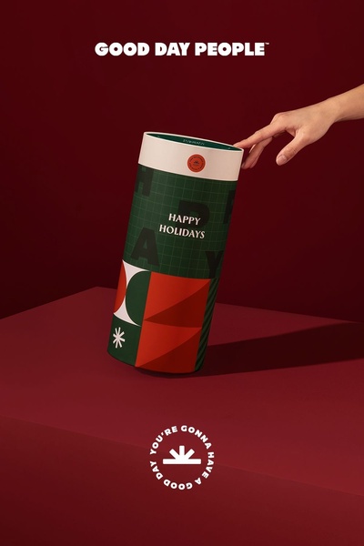 A high-end, minimalist presentation of a cylindrical gift box against a deep red background, focusing on refined branding and seasonal holiday messaging.