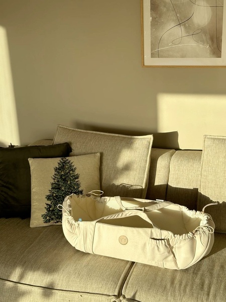 A high-quality, sun-drenched product photo of a baby nest resting on a couch, featuring a subtle Christmas tree decorative pillow and minimalist wall art.
