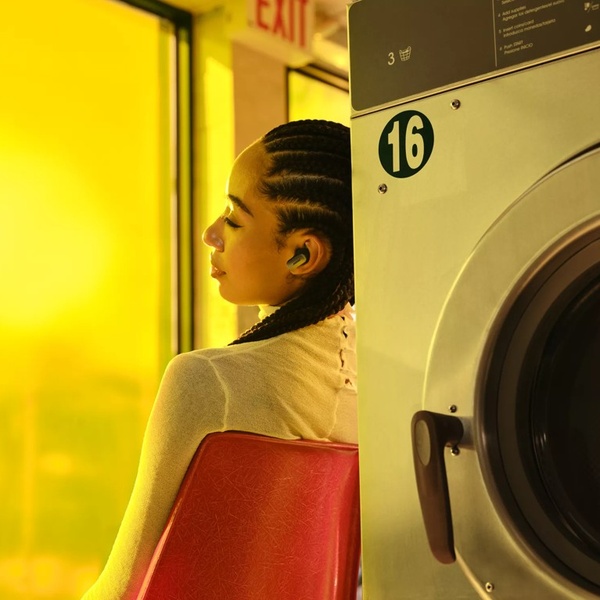 A close-up shot of a person wearing sleek wireless earbuds, leaning against a laundry machine in a warm, dimly lit room.