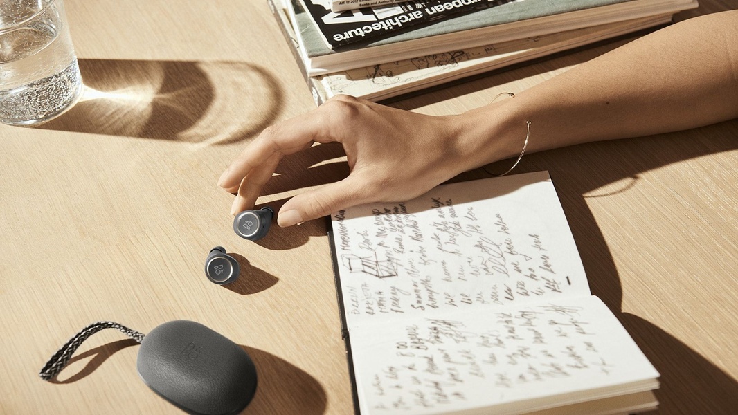 A high-end lifestyle shot featuring wireless earbuds and a notebook on a wooden desk, emphasizing a clean, premium aesthetic.